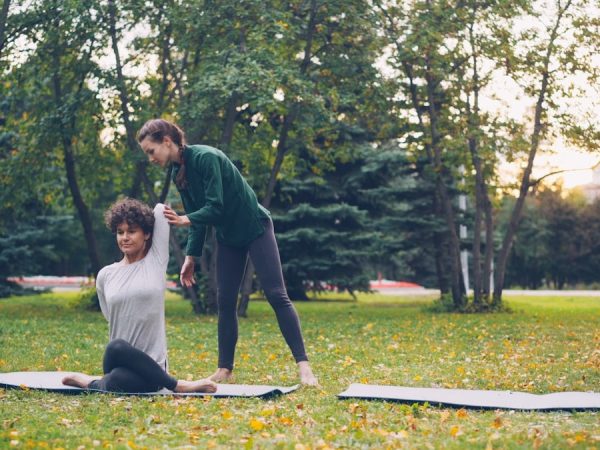 Diligent yoga student is learning Cow Face position sitting on mat and stretching arms while experienced teacher is helping her, girls are talking and laughing at outdoor ptactice.