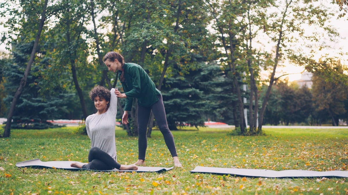 Diligent yoga student is learning Cow Face position sitting on mat and stretching arms while experienced teacher is helping her, girls are talking and laughing at outdoor ptactice.